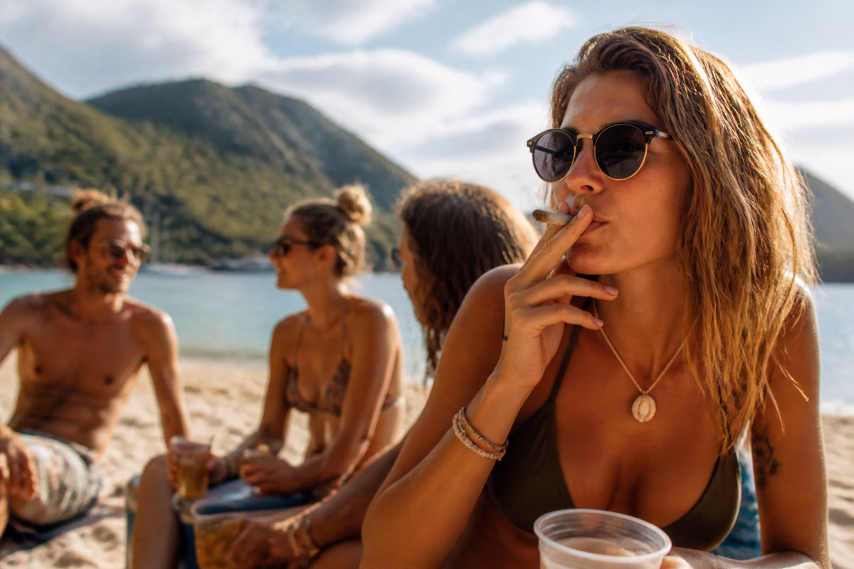 Femeie cu ochelari de soare fumând un joint pe o plajă tropicală, cu prieteni cu băuturi în fundal