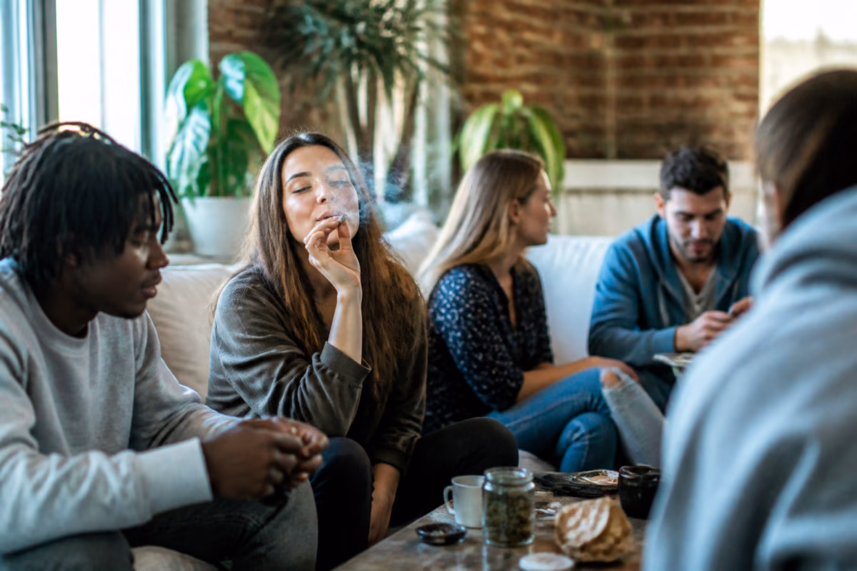 Tineri adulți pe o canapea, o femeie fumează un joint, pe masă un borcan cu canabis