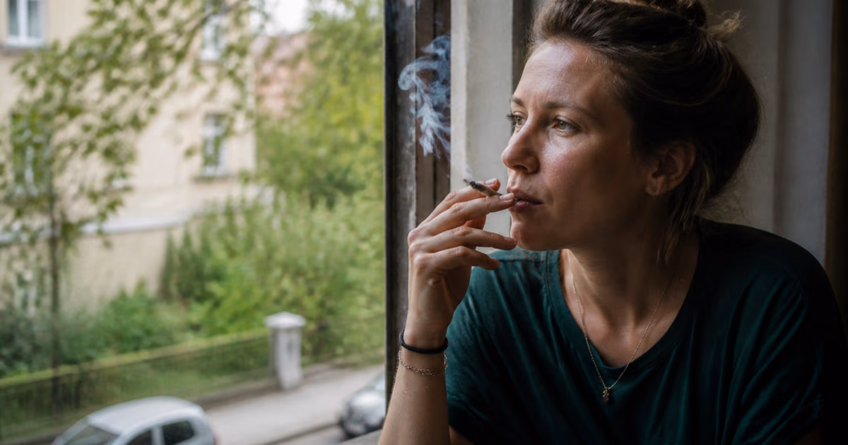 Femeie fumând un joint lângă fereastră și privind o stradă cu copaci